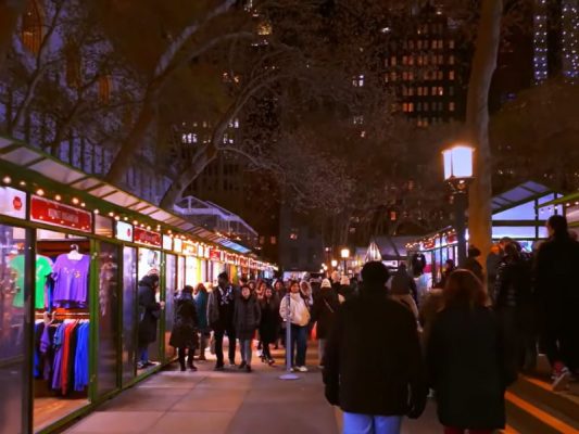 Keramaian pengunjung di Bryant Park Winter Village pada malam hari dengan deretan kios belanja yang terang